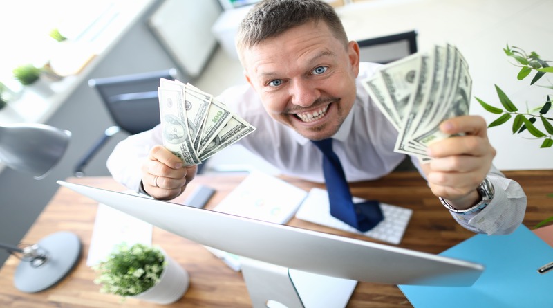 Homem sorridente segurando um maço de dinheiro na frente de um laptop aberto, demonstrando o conceito de ganhos rápidos online.
