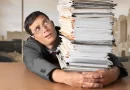 Homem sobrecarregado com pilha de documentos, simbolizando a exploração no trabalho moderno.